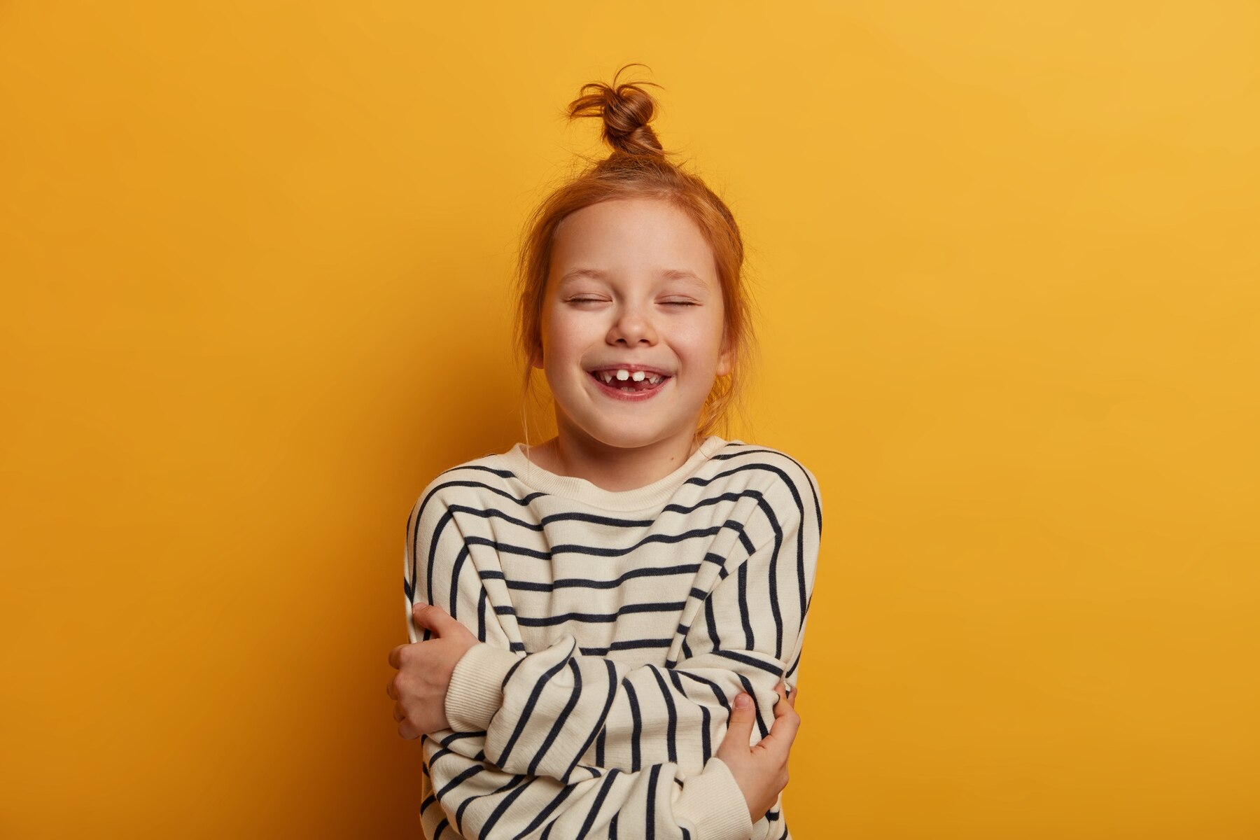 dealsginger child embraces herself expresses self love closes eyes from pleasure wears striped jumper poses against yellow wall feels good shows white teeth full excitement273609 42096